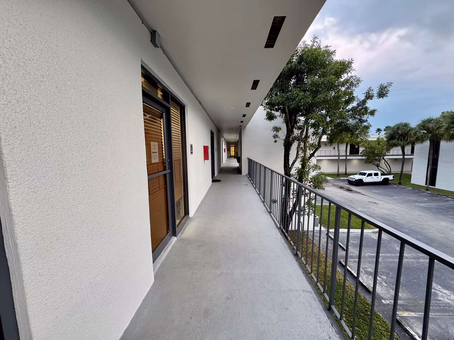Long outdoor corridor along a white building with doors, railing, and a nearby parking lot.