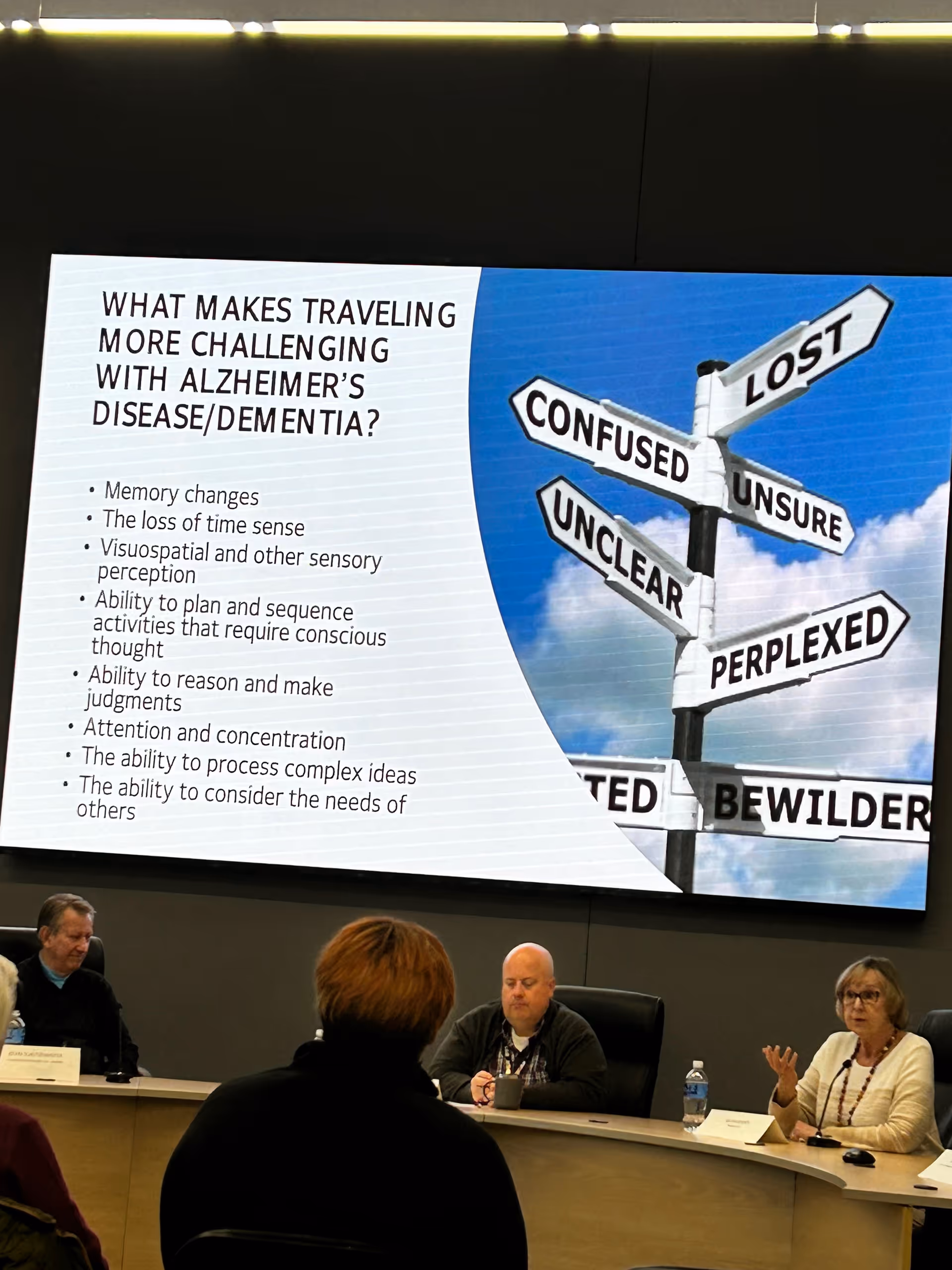 Panelists seated around a curved conference table as a large presentation screen displays dementia education.