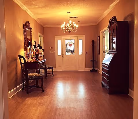 A warmly lit hallway with wooden flooring and beige walls. On the left side, there is a wooden console table with decorative items and a mirror above it, accompanied by two chairs. On the right side, there is a tall wooden cabinet. At the end of the hallway is a white front door with glass panels and a decorative wreath. A chandelier hangs from the ceiling.