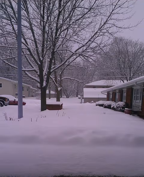 Snow-covered outdoor view of a facility's front area with trees, benches, and buildings.