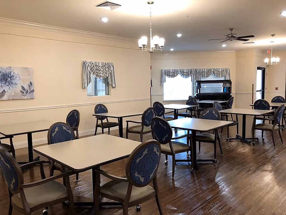A dining room with multiple square tables and blue cushioned chairs arranged neatly on a wooden floor. The room has cream-colored walls with decorative molding, windows with striped valances, ceiling lights, and a ceiling fan. A piano is visible near the windows in the background.