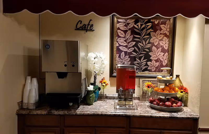 A small cafe counter with a beverage dispenser, a water machine, stacks of disposable cups, and a three-tiered tray holding apples, oranges, and bananas. The counter is decorated with artificial flowers and a framed floral artwork on the wall behind it. A sign above the water machine reads 'Cafe'.