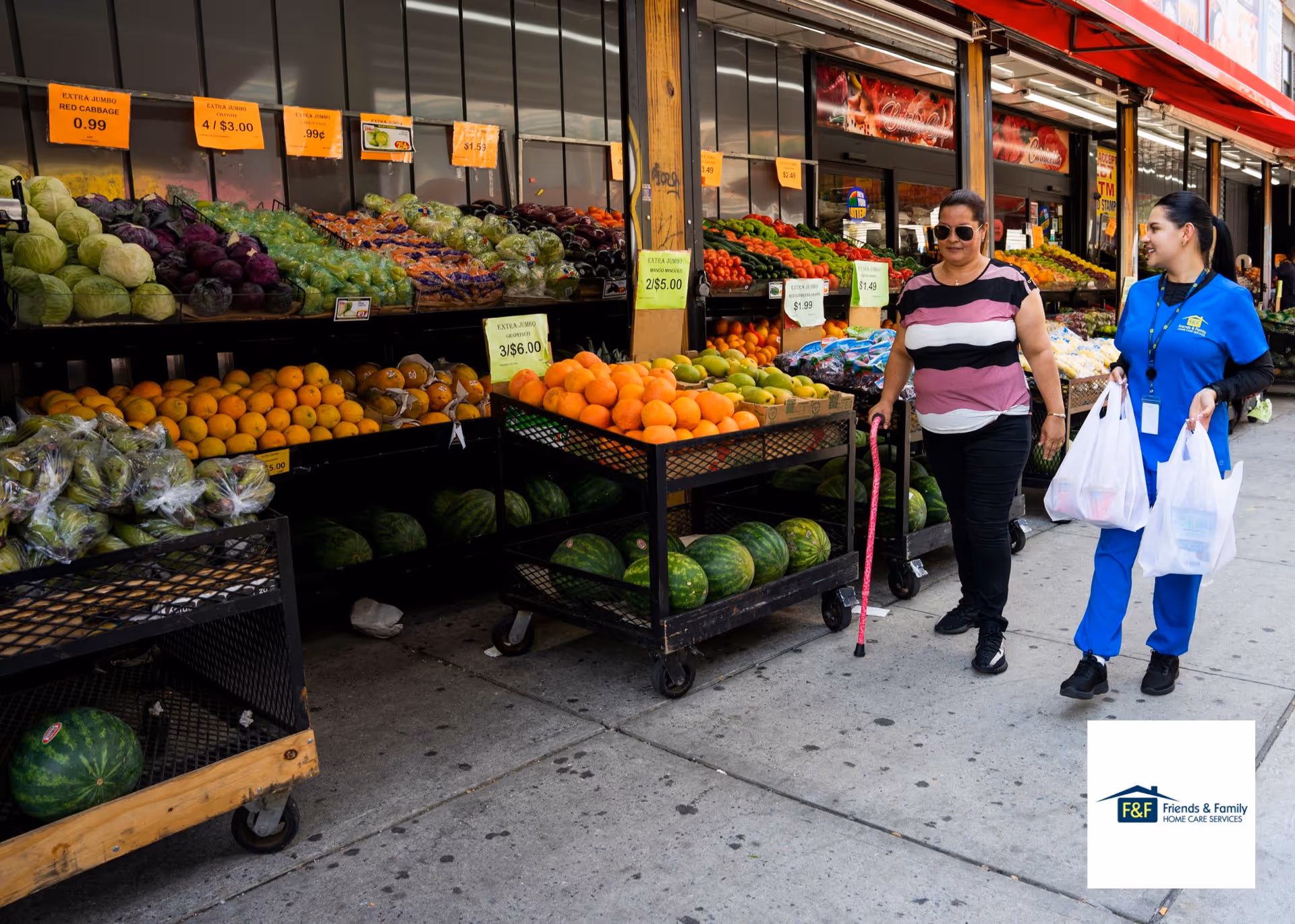Two women with shopping bags browse a sidewalk fruit stand outside a market, featuring colorful produce.