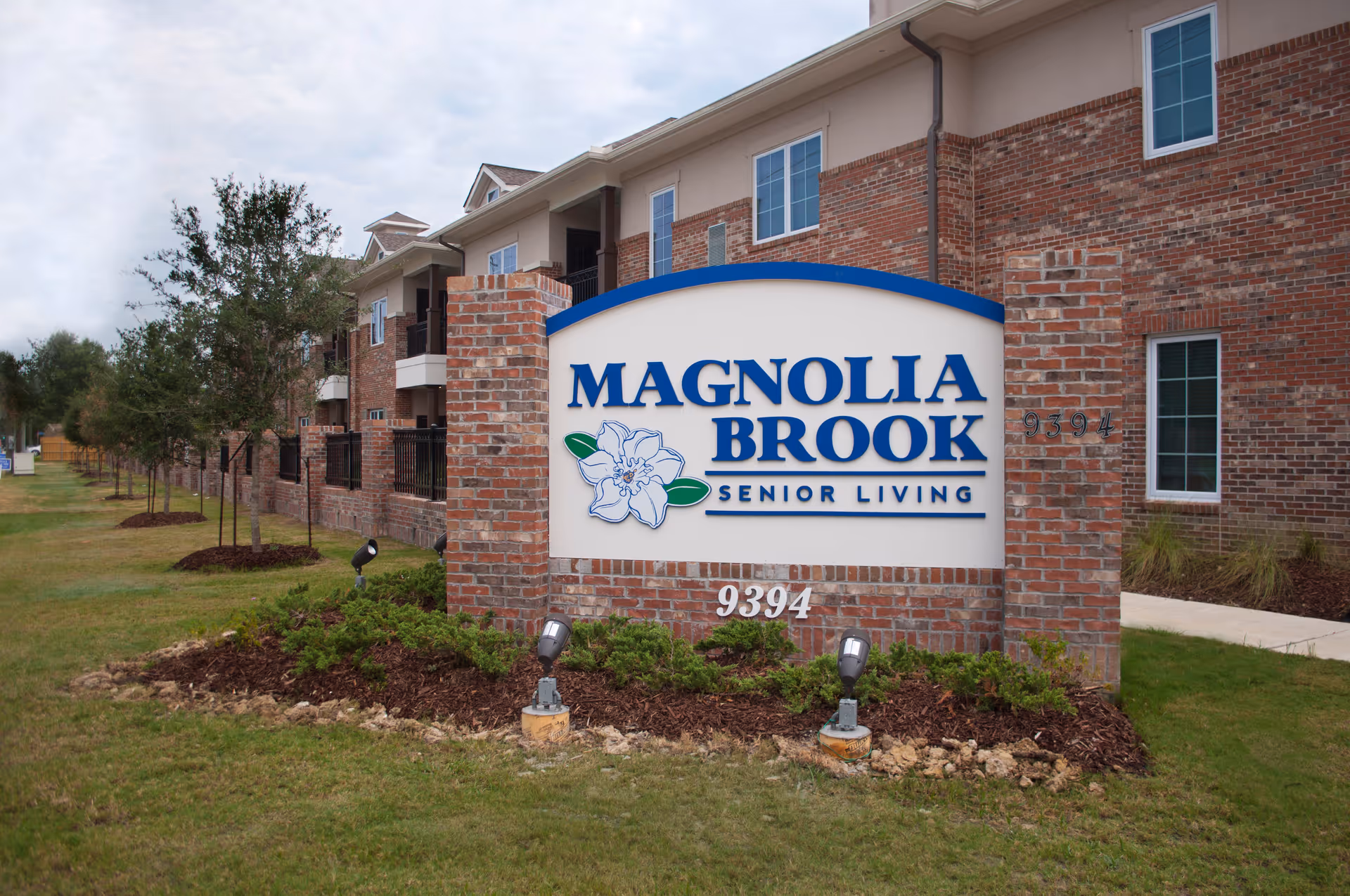 Exterior view of a senior living facility with a large brick sign in the foreground that reads 'MAGNOLIA BROOK SENIOR LIVING' with a magnolia flower graphic. The building is made of red brick with white trim and multiple windows. There is a grassy area with small trees and landscaping around the sign.