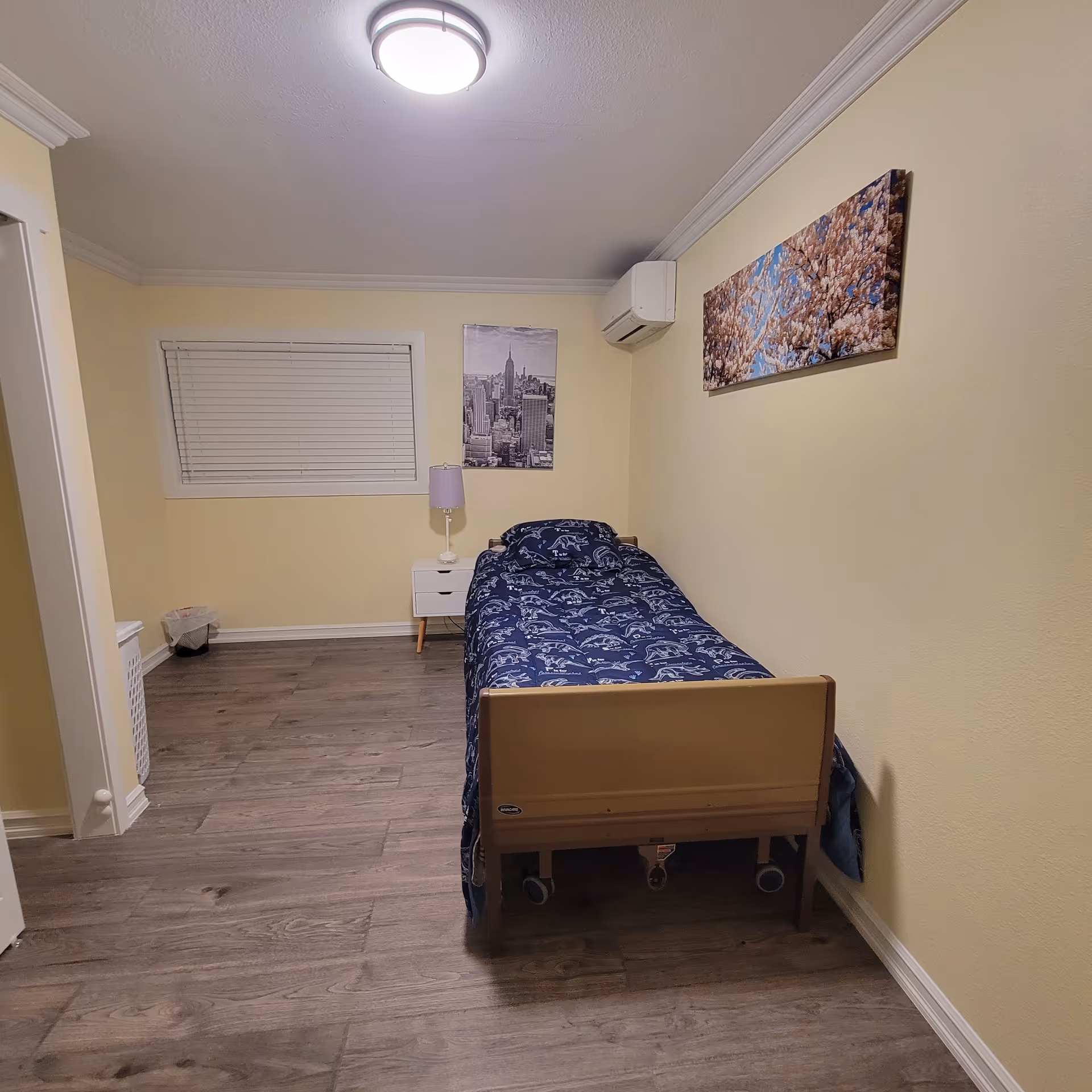 A small bedroom with a single bed covered with a blue patterned blanket. There is a white nightstand with a purple lamp next to the bed. The walls are painted light yellow and decorated with two framed pictures, one of a cityscape and the other of blooming flowers. The floor is wooden, and there is a window with closed blinds on the left wall. An air conditioning unit is mounted on the wall above the bed.
