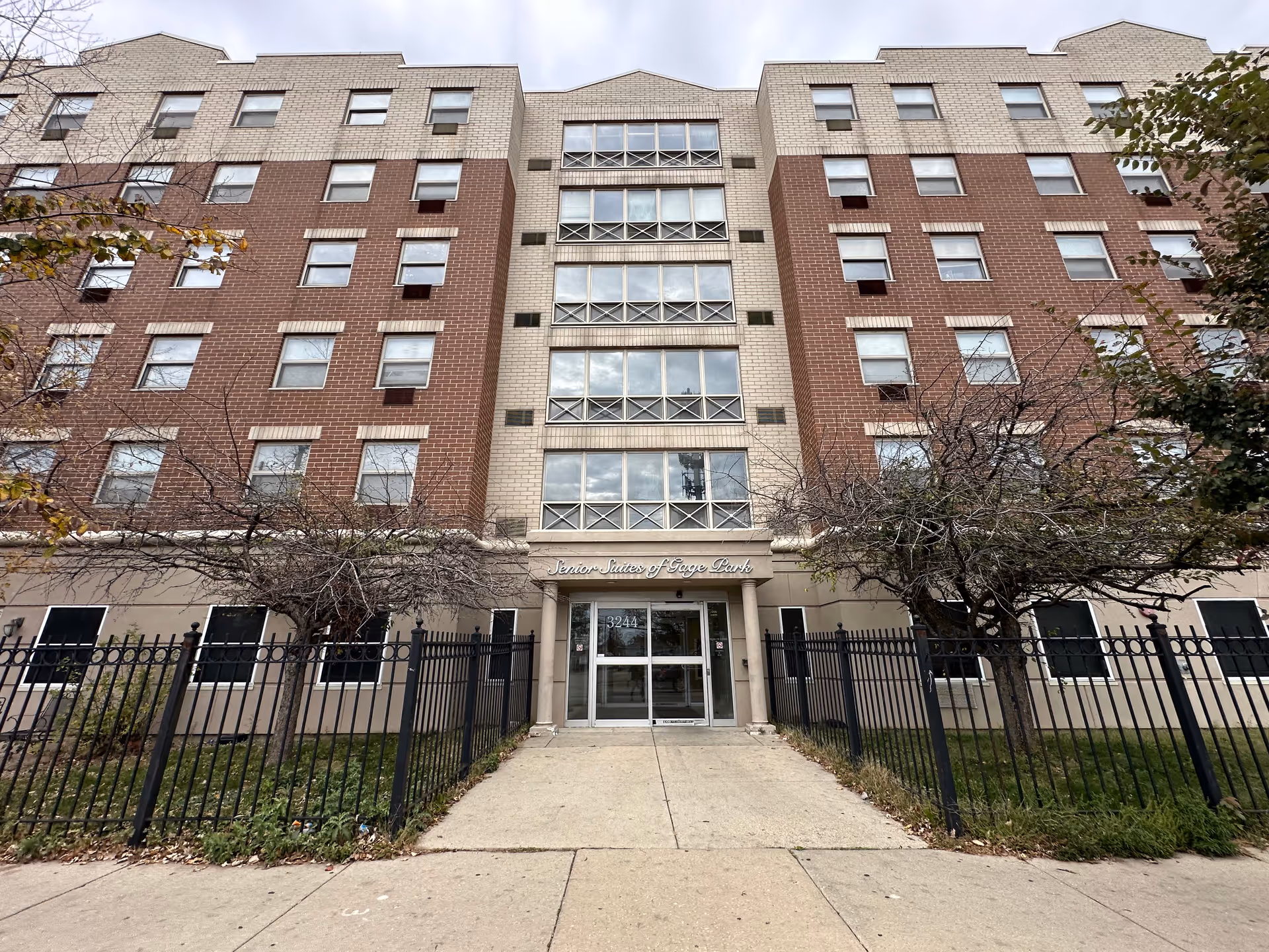 Front façade of a multi-story brick-and-stucco senior living building with a gated entrance and a sign reading 'Senior Suites of Gage Park' above the doors.