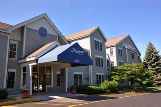 Front entrance of a light-gray, multi-story residential building with a blue awning that reads 'Lake Square' and surrounding landscaping.