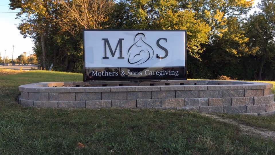 Outdoor sign for Mothers and Sons Caregiving displayed on a circular brick planter.