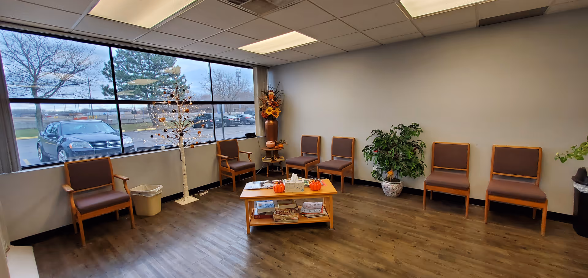 A quiet waiting area with brown chairs, a central coffee table, plants, pumpkins, and large windows.