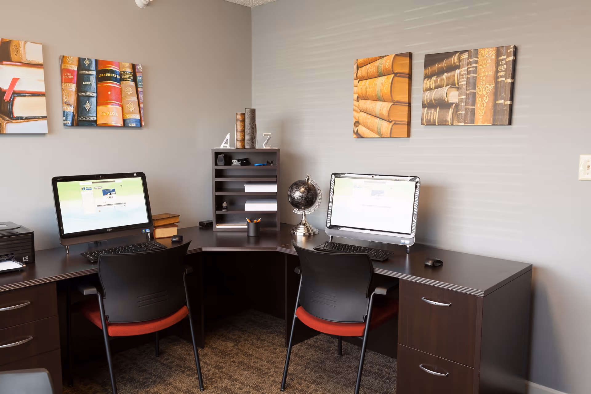 A small office area with two computer workstations on a dark wood L-shaped desk. Each workstation has a computer monitor, keyboard, and mouse. Two black chairs with red cushions are positioned at the desks. On the desk are a small globe, a pencil holder with pencils, and a small organizer with shelves. The walls are painted light gray and decorated with three framed pictures of old books.