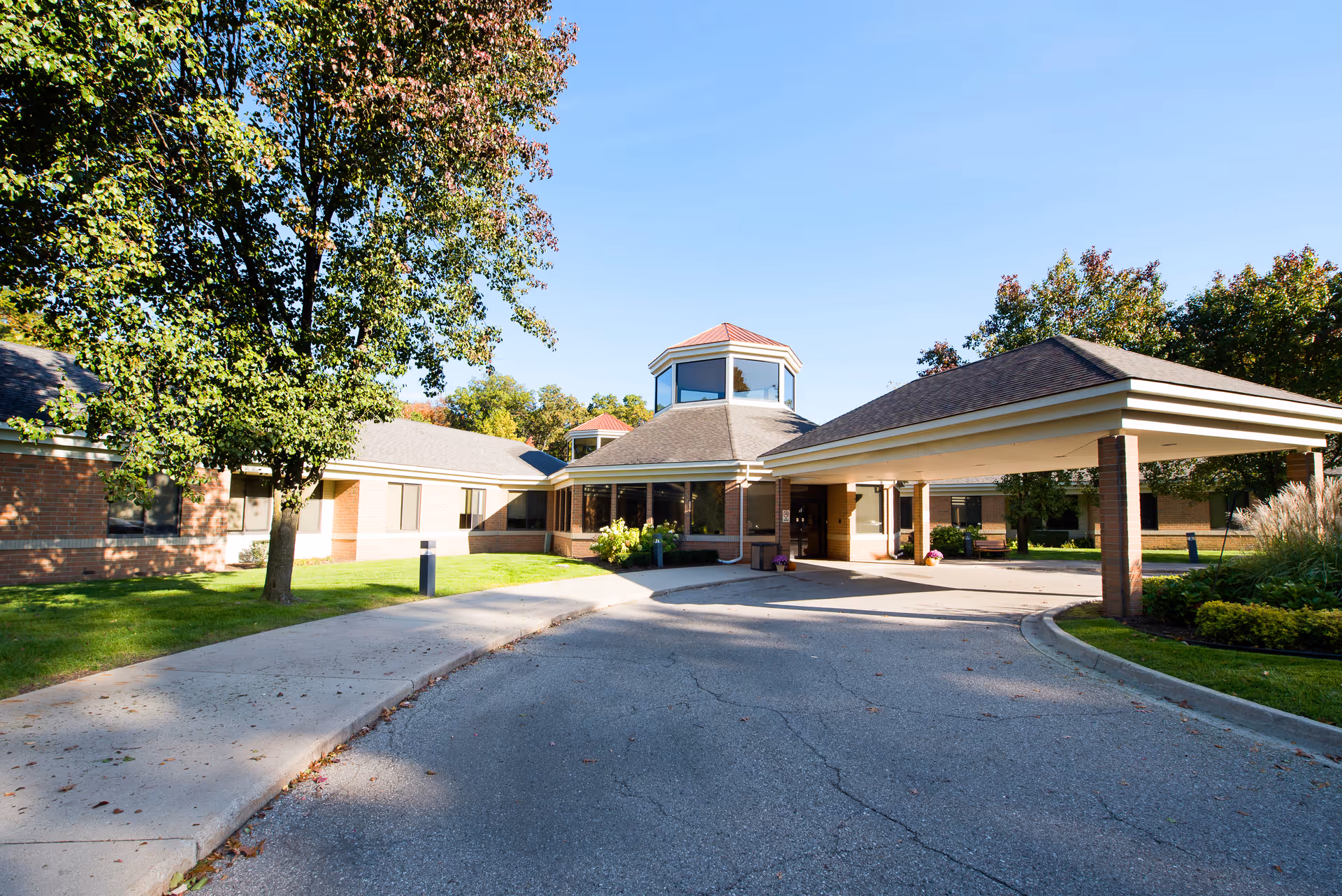 Exterior view of Wellspring Lutheran Services - Livonia Woods Campus building entrance with a covered drop-off area, surrounded by trees and greenery under a clear blue sky.
