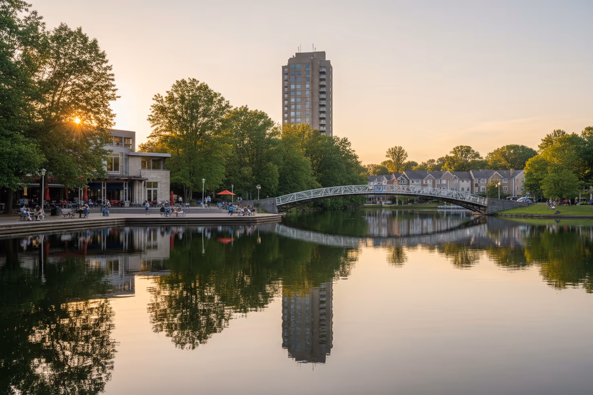 Sunset-lit lakefront with trees, a modern building, and a curved footbridge reflected on calm water.