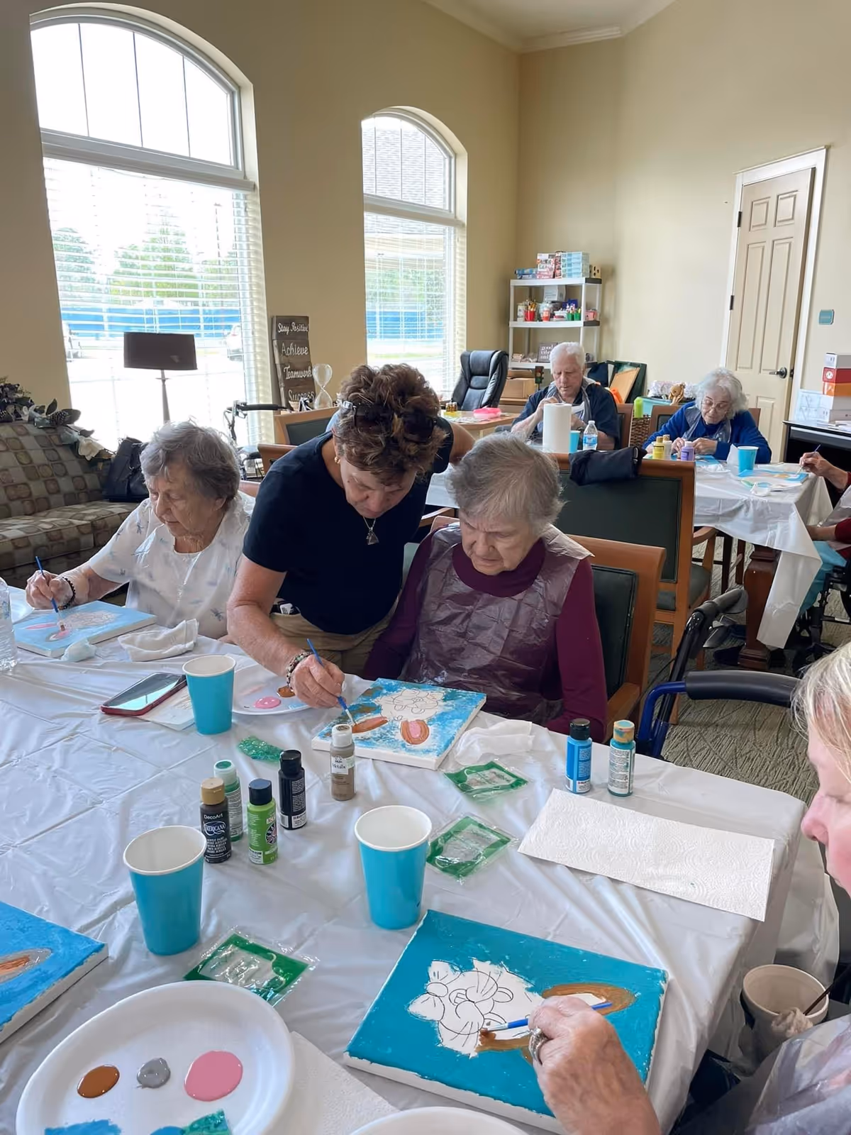 A group of elderly individuals seated around tables in a well-lit room, engaged in a painting activity with canvases, paint bottles, and brushes. A caregiver or instructor is assisting one of the elderly women with her painting. The room has large windows, comfortable chairs, and shelves with supplies in the background.