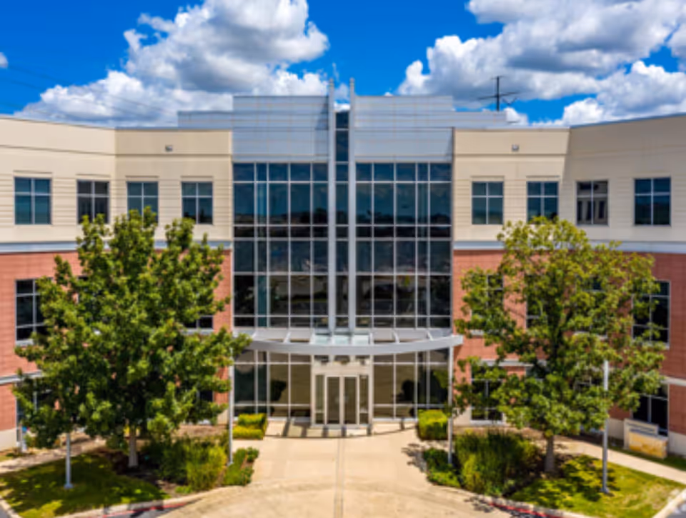 Modern healthcare facility exterior with glass entrance, beige walls, and green trees.