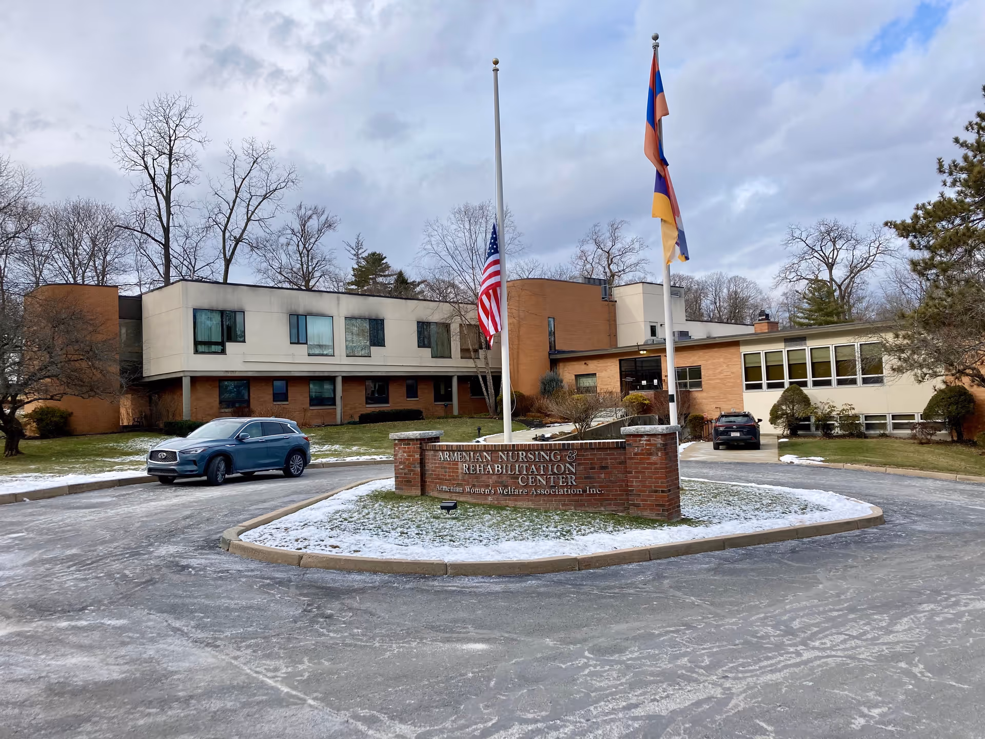 Front view of the Armenian Nursing & Rehabilitation Center building with a circular driveway, flagpoles, and light snow on the ground.