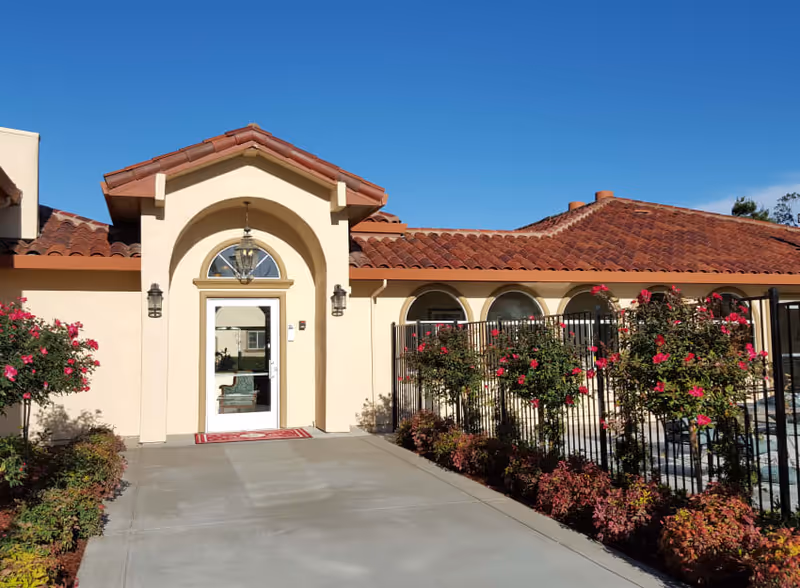 Exterior view of a building with beige walls and a red tiled roof under a clear blue sky. The entrance features an arched doorway with a glass door and a hanging lantern light fixture. There are flowering bushes with pink flowers along a black metal fence on the right side of the walkway leading to the entrance.