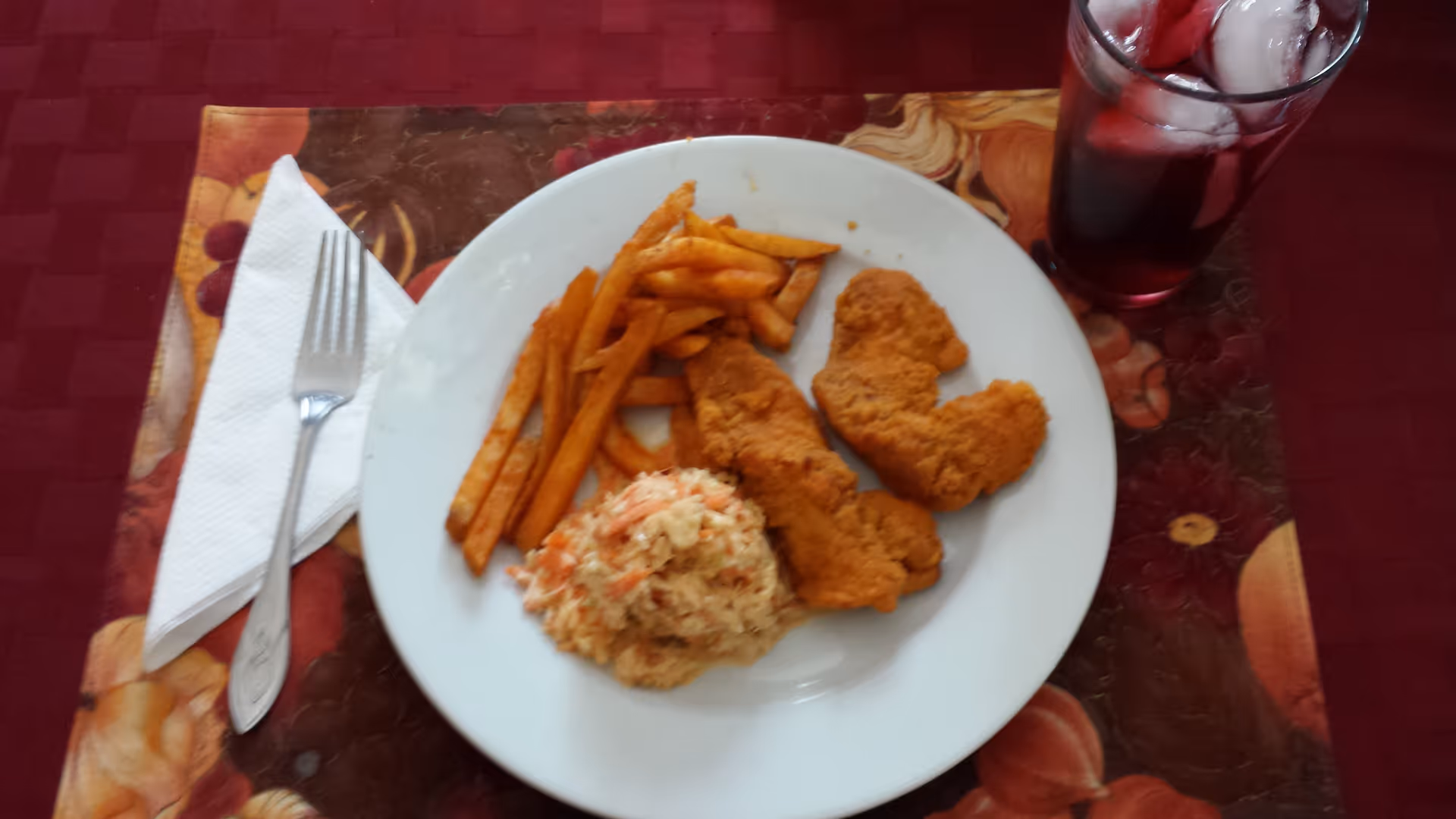 A white plate with fried chicken strips, French fries, and coleslaw on a decorative placemat. To the left of the plate is a fork on a folded white napkin. To the right is a glass of iced dark-colored beverage.