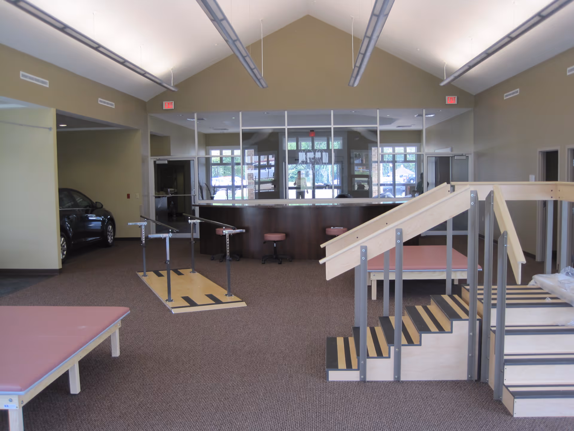 Interior view of a rehabilitation or physical therapy room with parallel bars for walking exercises, a set of wooden stairs with handrails for mobility training, padded therapy tables, and a reception desk in the background. The room has high ceilings with hanging lights and large windows behind the reception area.