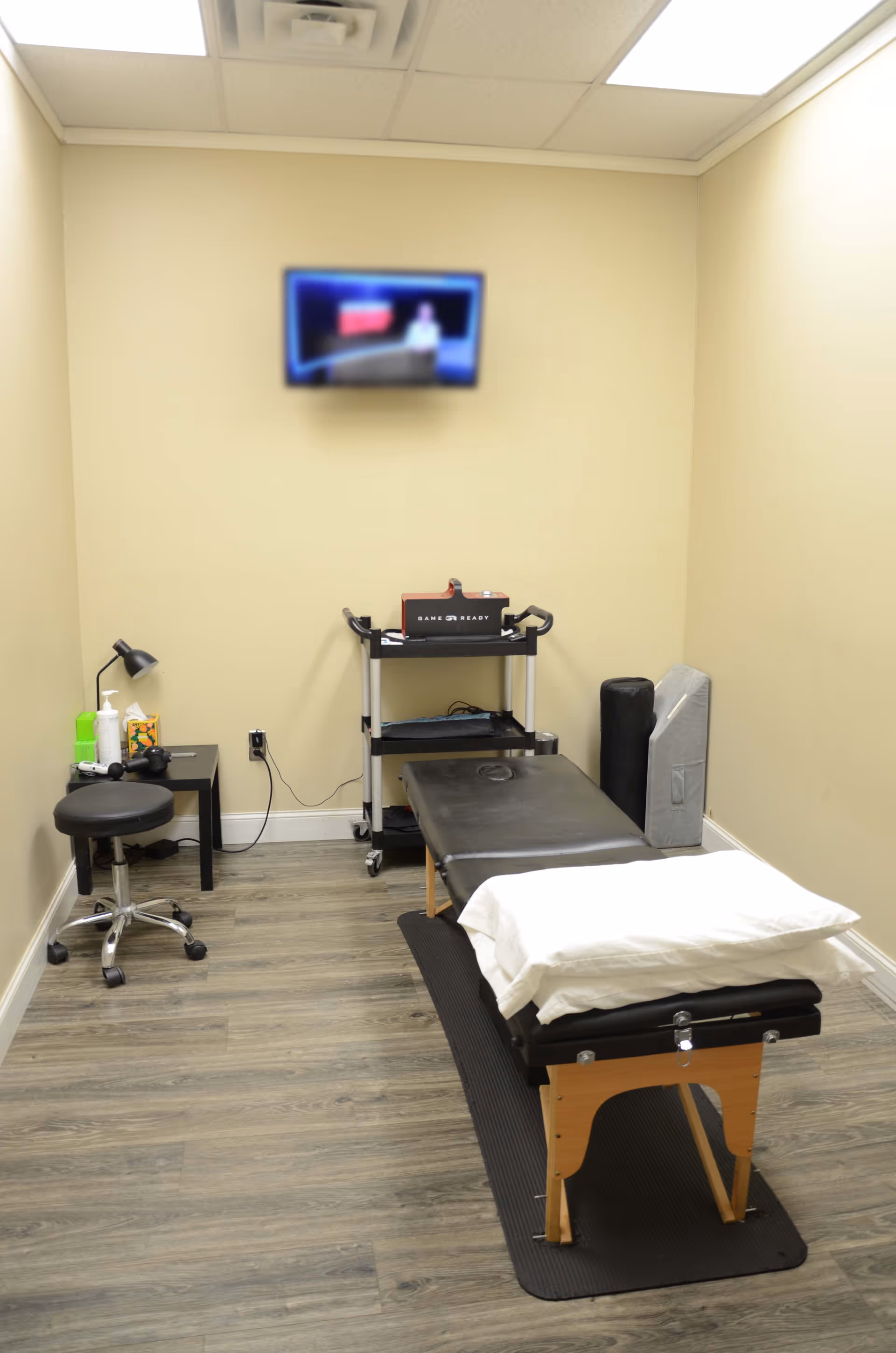 A compact therapy room with an examination table, rolling cart, stool, and wall-mounted monitor.