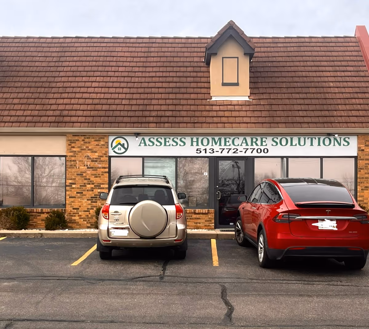 Front view of a beige brick building with Assess Homecare Solutions sign and parked cars outside.