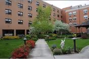 Brick senior living building with a central courtyard, walkway, trees, shrubs, and a white garden statue.