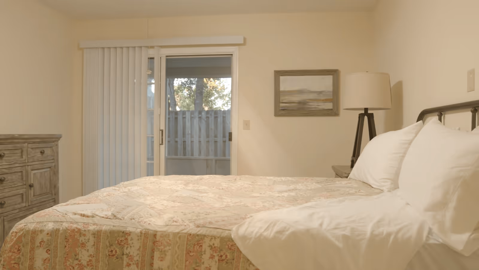 A cozy bedroom with a floral patterned bedspread on a bed with white pillows. There is a wooden dresser on the left side, a sliding glass door with vertical blinds leading outside, a framed landscape painting on the wall, and a floor lamp next to the bed.