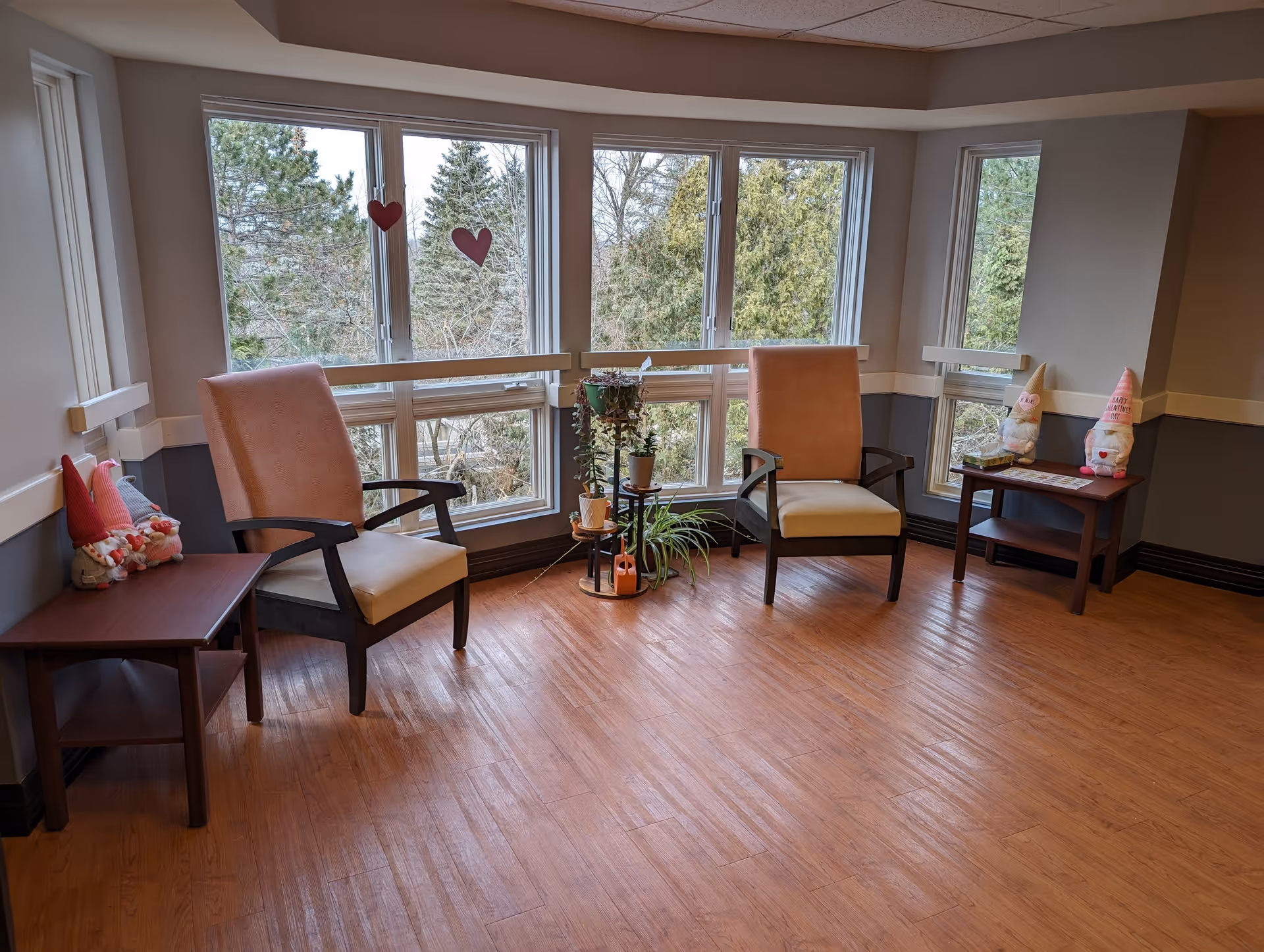 Bright senior living lounge with two beige chairs, small tables, plants, and heart decorations on windows.