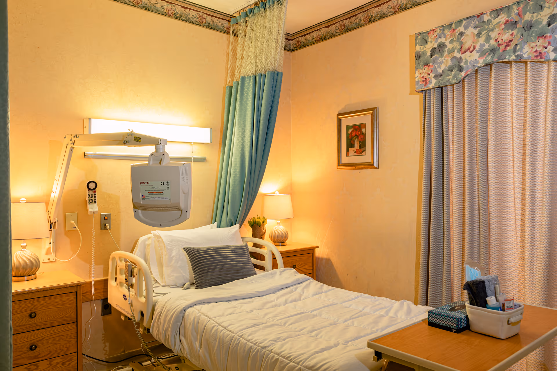 Hospital-style patient room with a bed, privacy curtains, bedside tables, and a wall-mounted medical device.