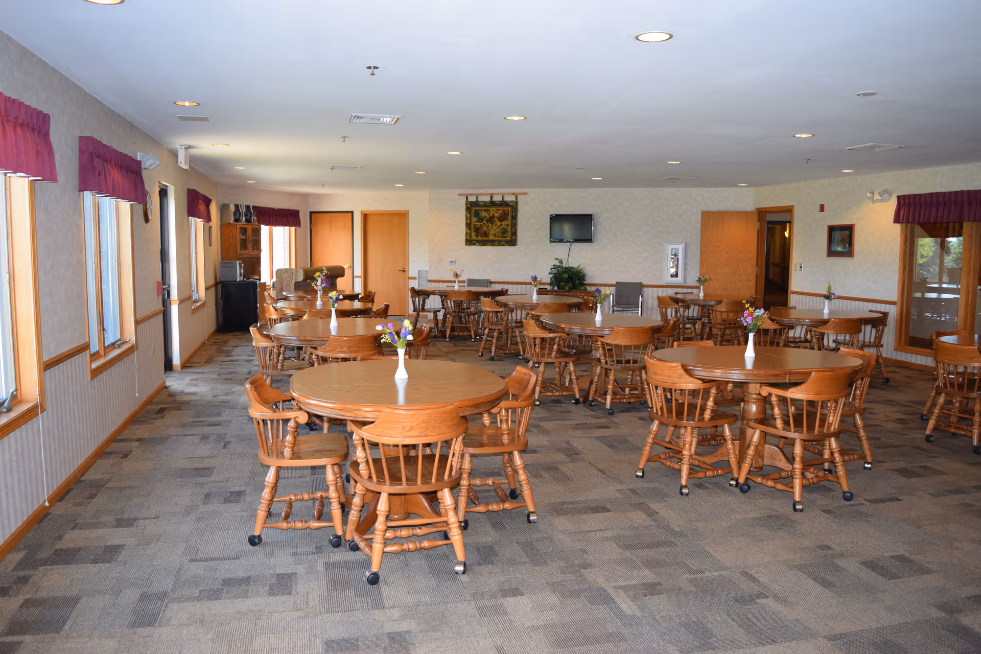Spacious senior living dining room with round wooden tables and floral centerpieces.