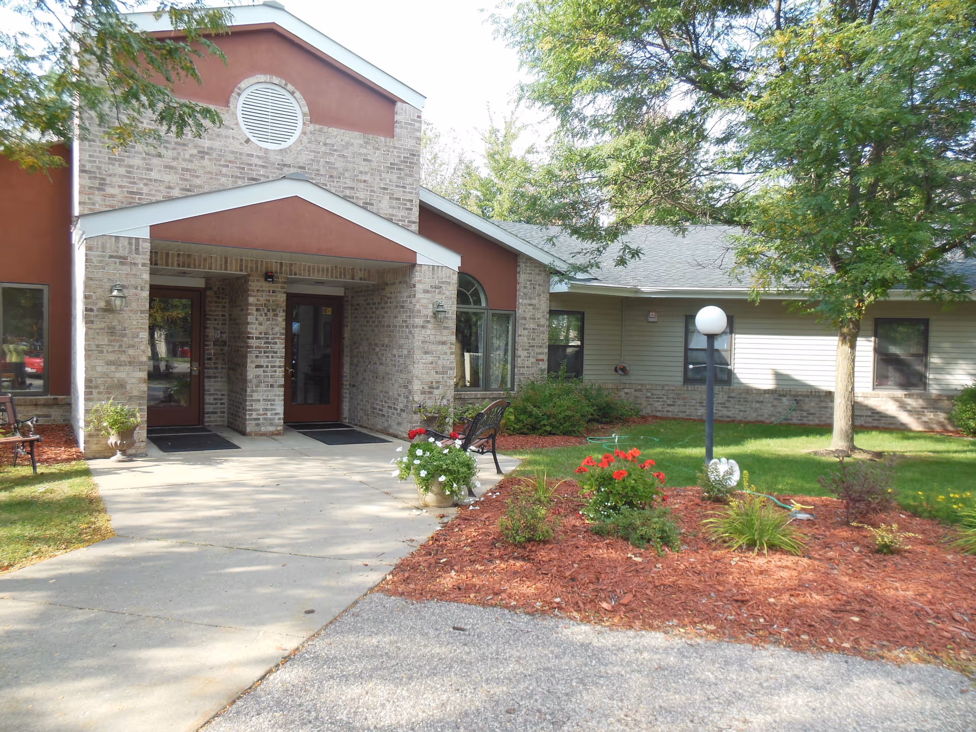 Exterior view of the entrance to Sylvan Crossings of Stevens Point, showing a brick and siding building with a covered entryway, a concrete walkway, landscaped garden beds with flowers and shrubs, a bench, and trees providing shade.