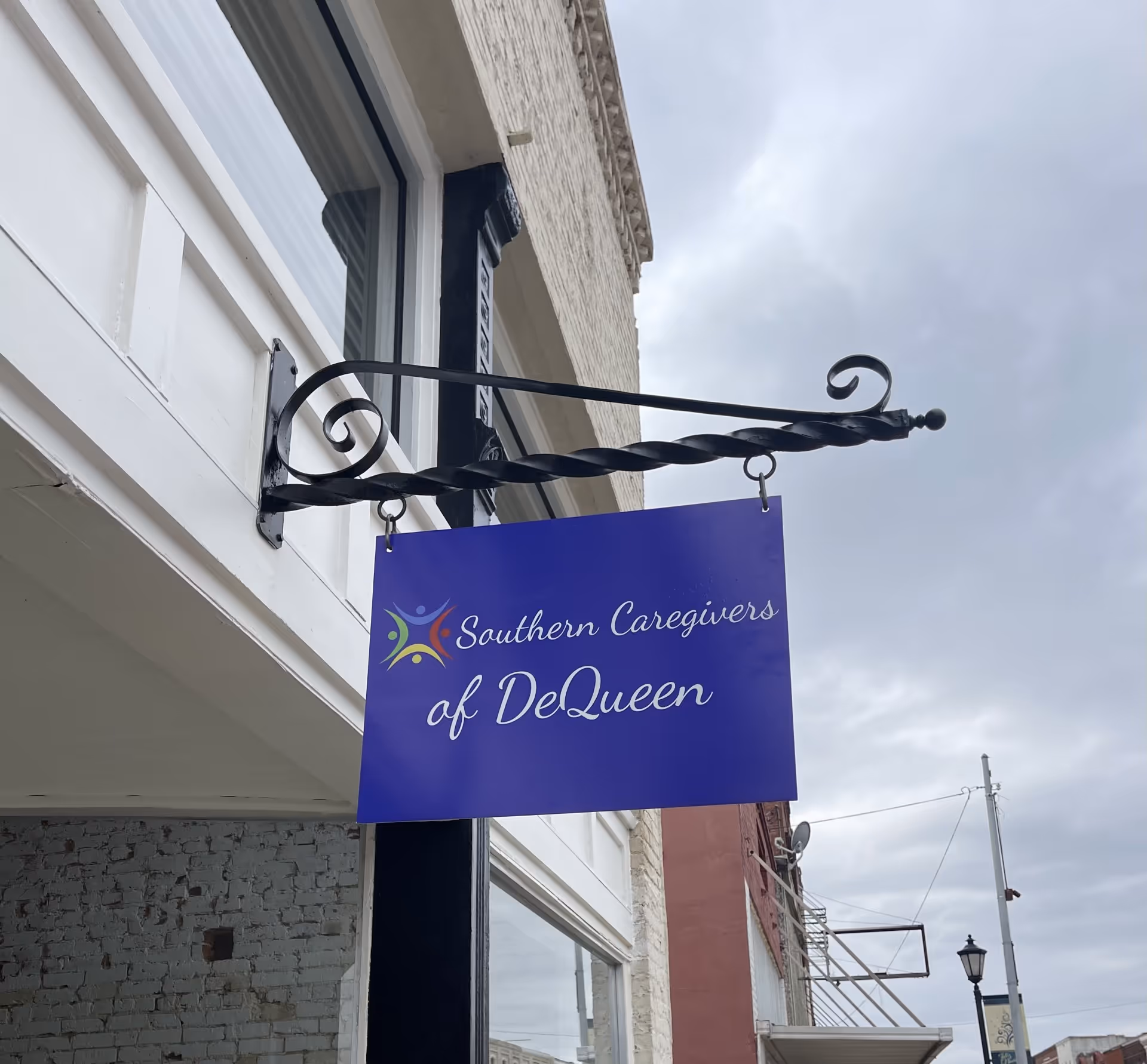 Blue sign reading Southern Caregivers of De Queen hangs from an ornate bracket on a building.