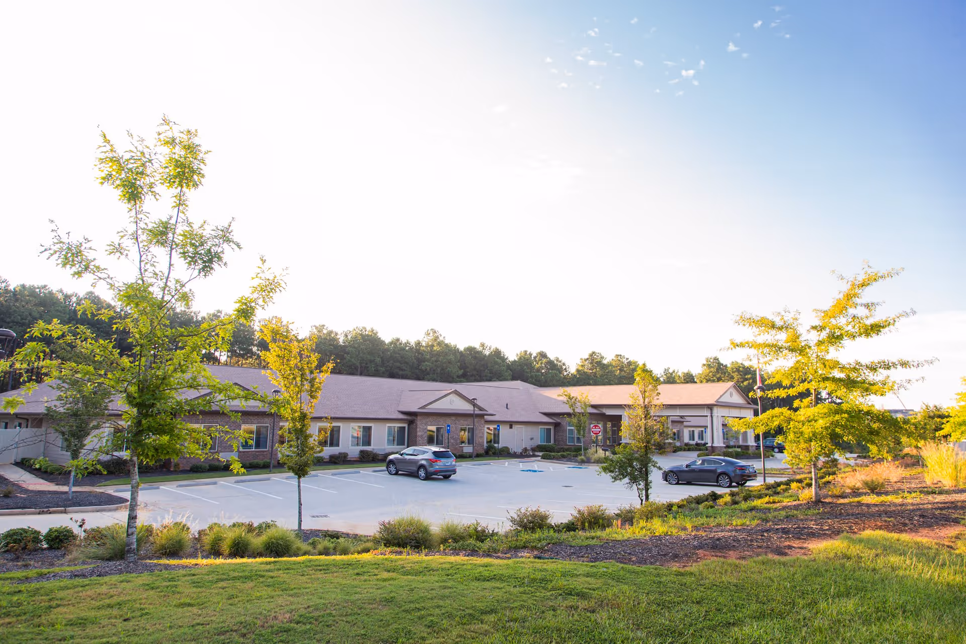 Exterior view of Marshall Pines Assisted Living & Memory Care building with a parking lot in front, surrounded by small trees and landscaped greenery under a clear blue sky.