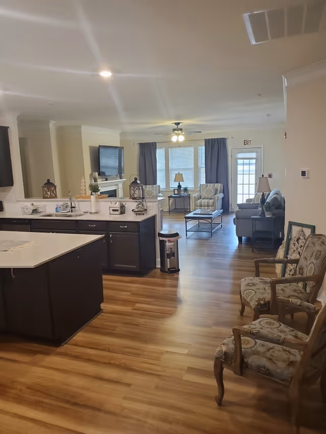 Interior view of a senior living facility showing a kitchen area with dark cabinets and a white countertop island, leading into a living room with armchairs, a sofa, a coffee table, and a TV mounted above a fireplace. The room has wooden flooring, large windows with curtains, and ceiling lights including a ceiling fan with lights.