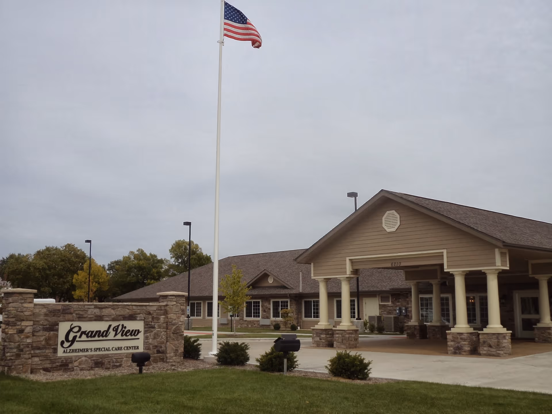 Exterior view of the Grand View Alzheimer's Special Care Center building with a covered entrance supported by columns, a stone sign with the facility name, an American flag on a tall flagpole, and some trees in the background under a cloudy sky.