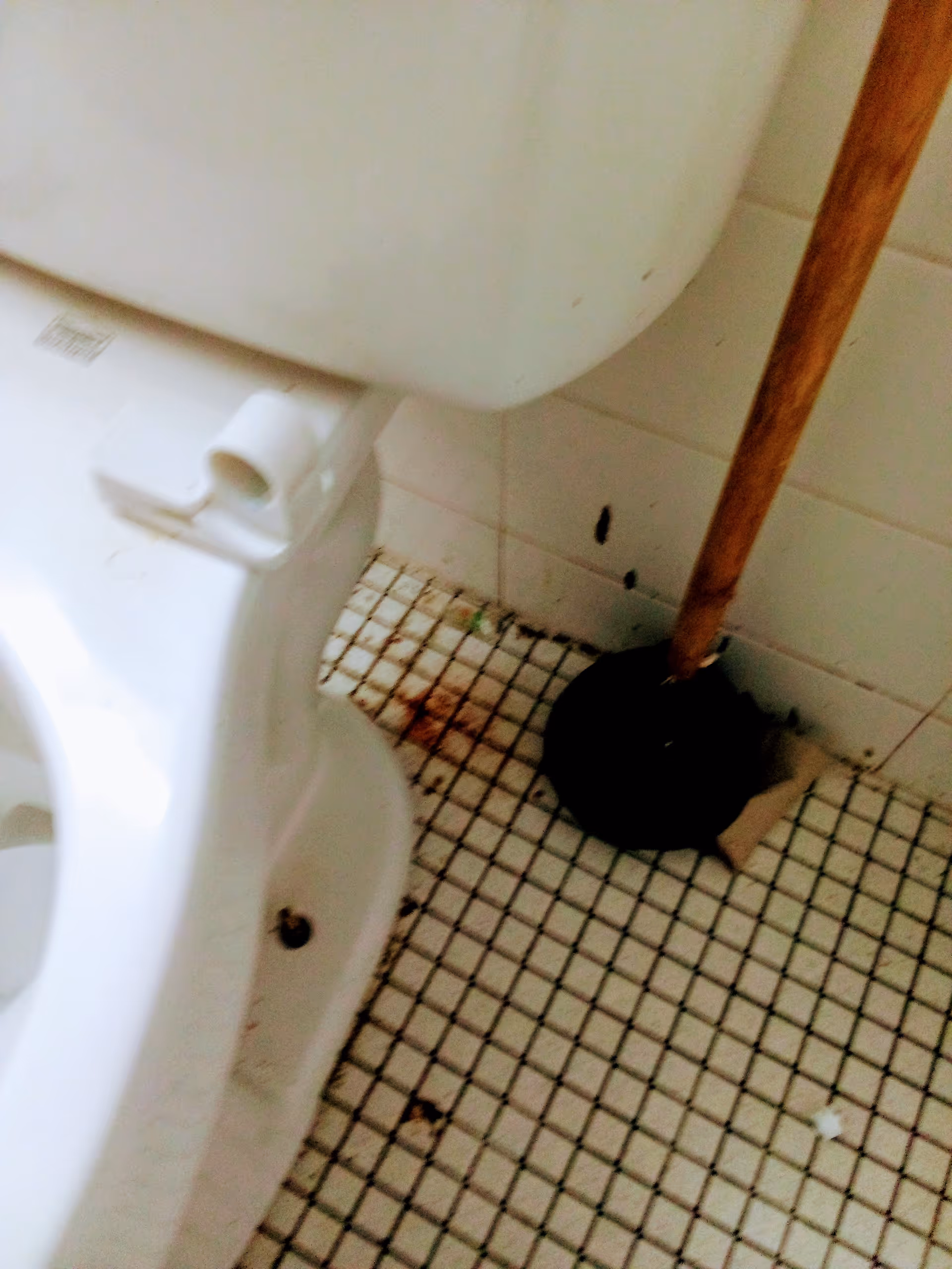 Toilet in a tiled bathroom with a mop leaning against the wall.