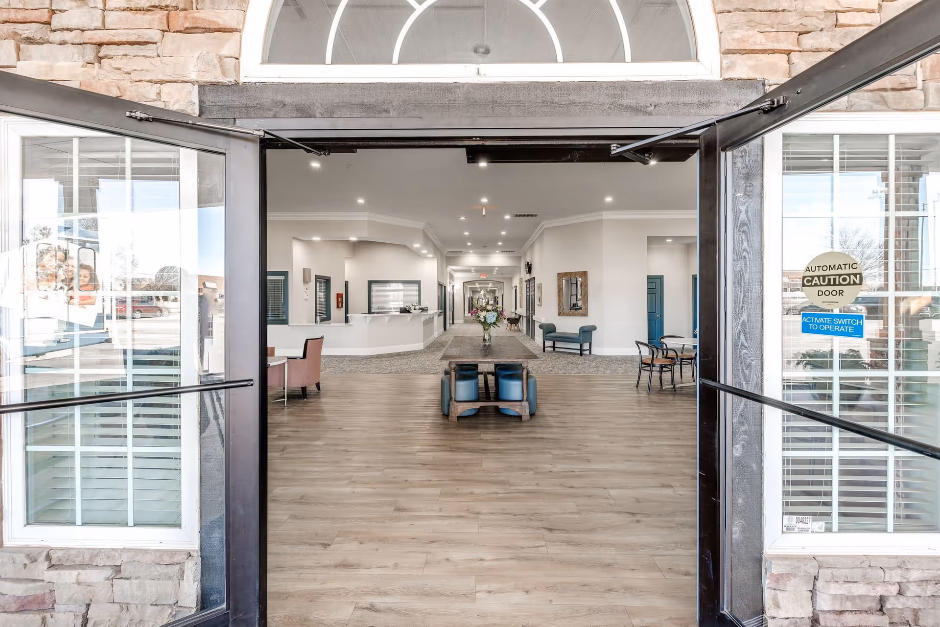 View through open double glass doors into a spacious senior living facility lobby with wood flooring, a long table with flower arrangements in the center, seating areas with chairs along the walls, and a reception desk on the left side. The walls are light-colored with several doorways and decorative mirrors.