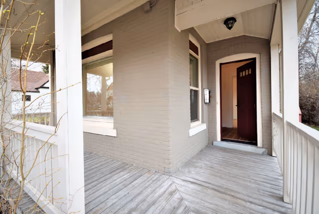 Wooden wraparound porch of a light brick building with an open front door.