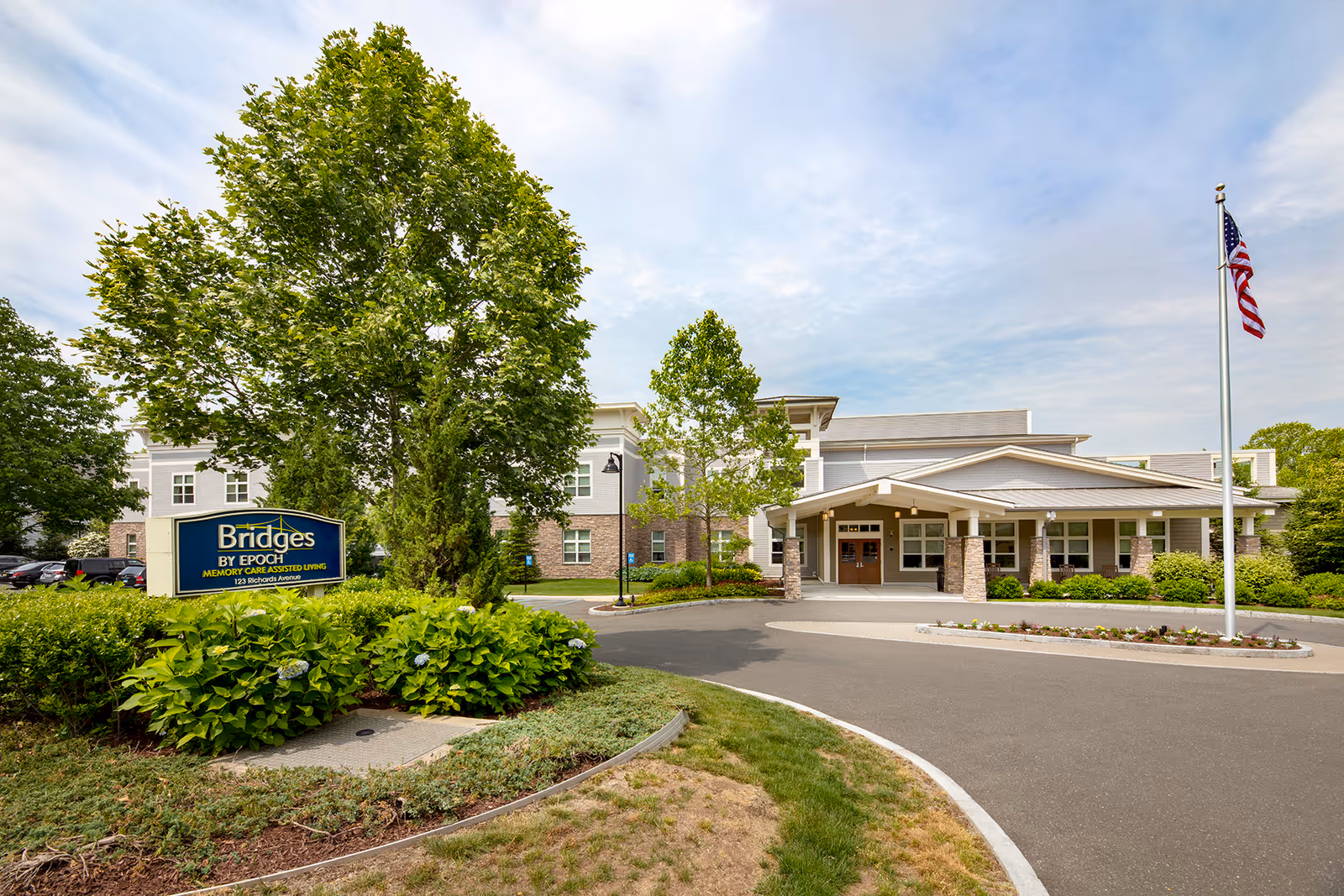 Front entrance of the Bridges by EPOCH assisted living building with a landscaped driveway, sign, and an American flag.