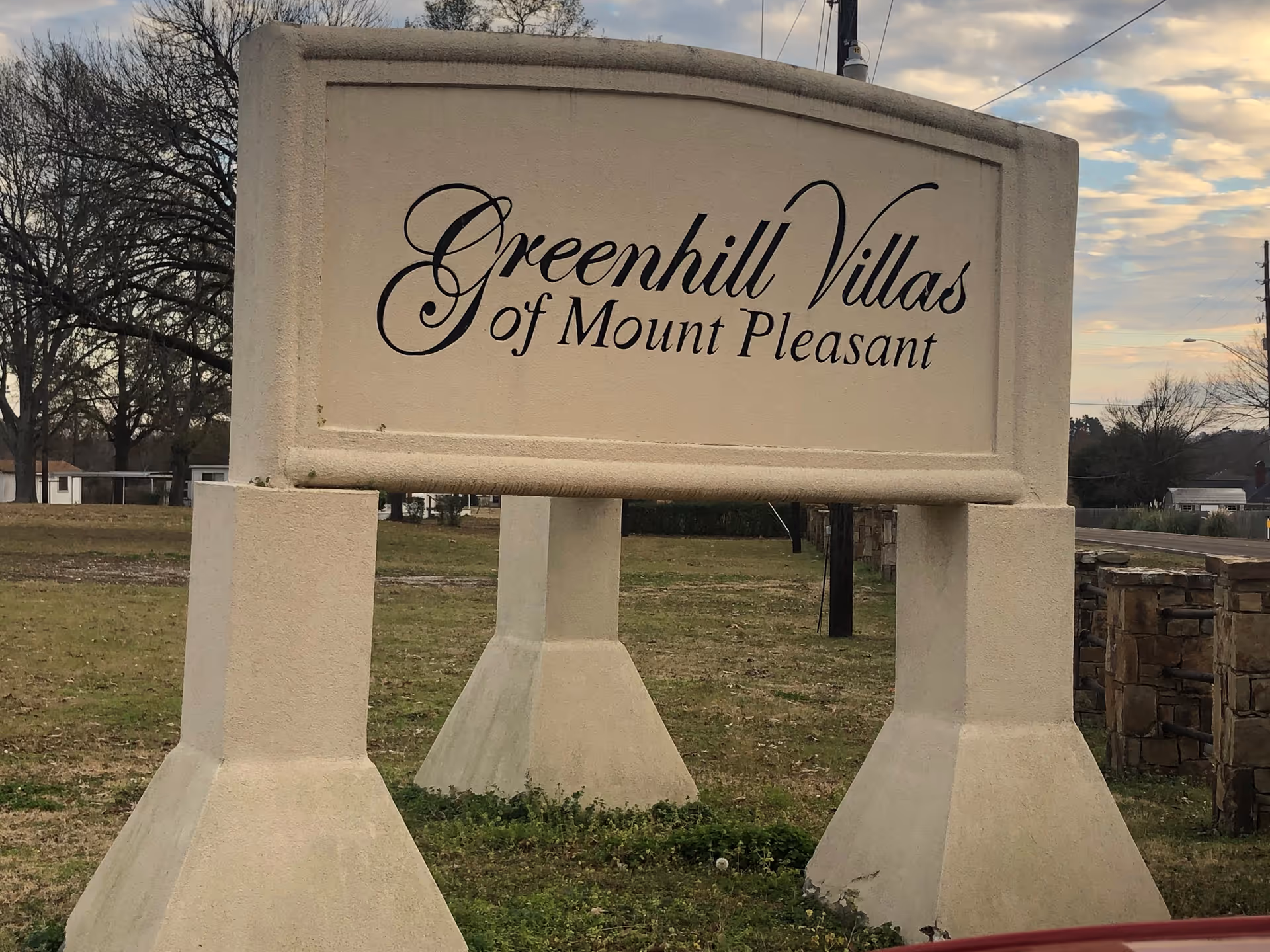 A large beige stone sign with black cursive text that reads 'Greenhill Villas of Mount Pleasant' situated on a grassy area with trees and a road in the background under a partly cloudy sky.