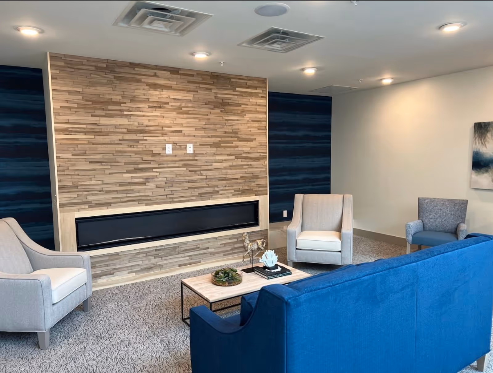 A modern living room with a blue sofa, two beige armchairs, and one blue armchair arranged around a rectangular coffee table. The back wall features a decorative wooden panel with a built-in electric fireplace. The room has recessed ceiling lights and a carpeted floor with a subtle pattern.