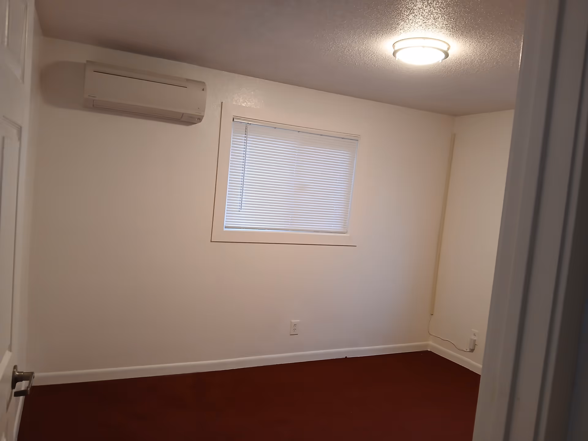Small empty bedroom with window, blinds, ceiling light, and wall-mounted AC unit.
