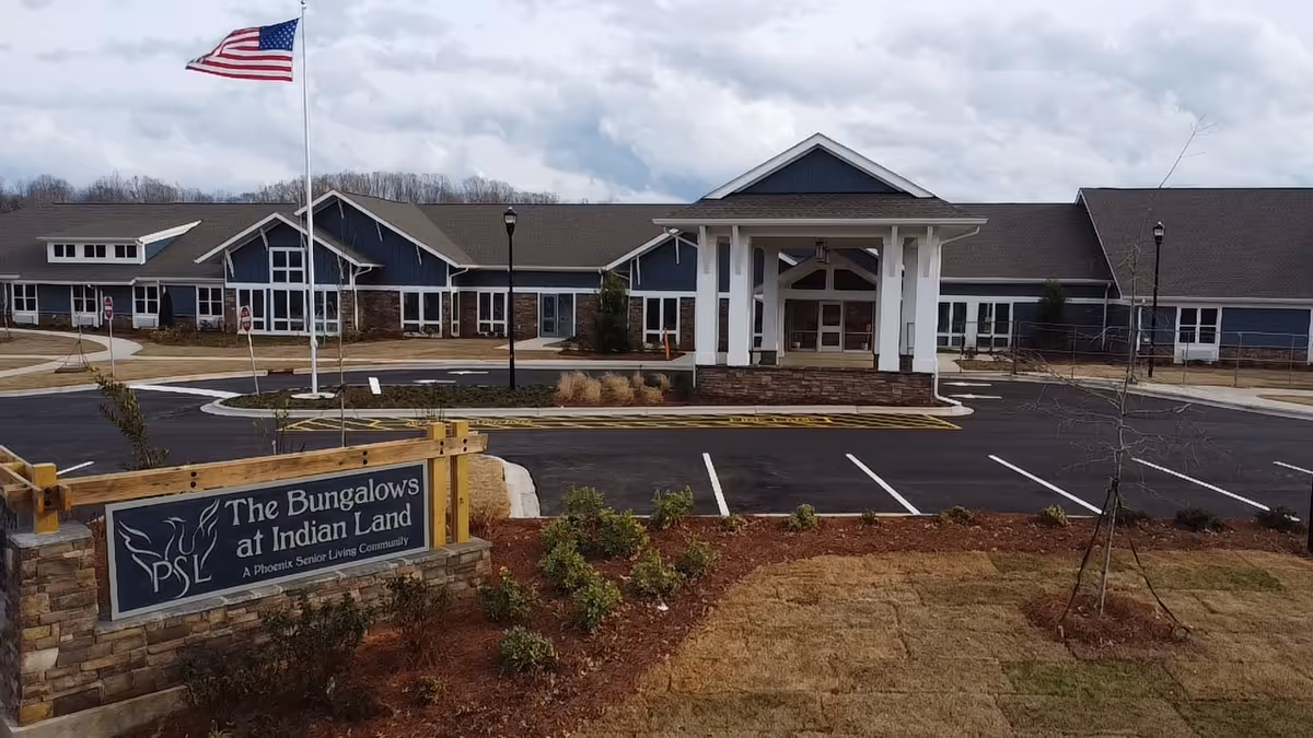 Exterior view of The Bungalows at Indian Land senior living facility with a large building, an American flag on a flagpole, a parking lot, and a landscaped area with bushes and grass.
