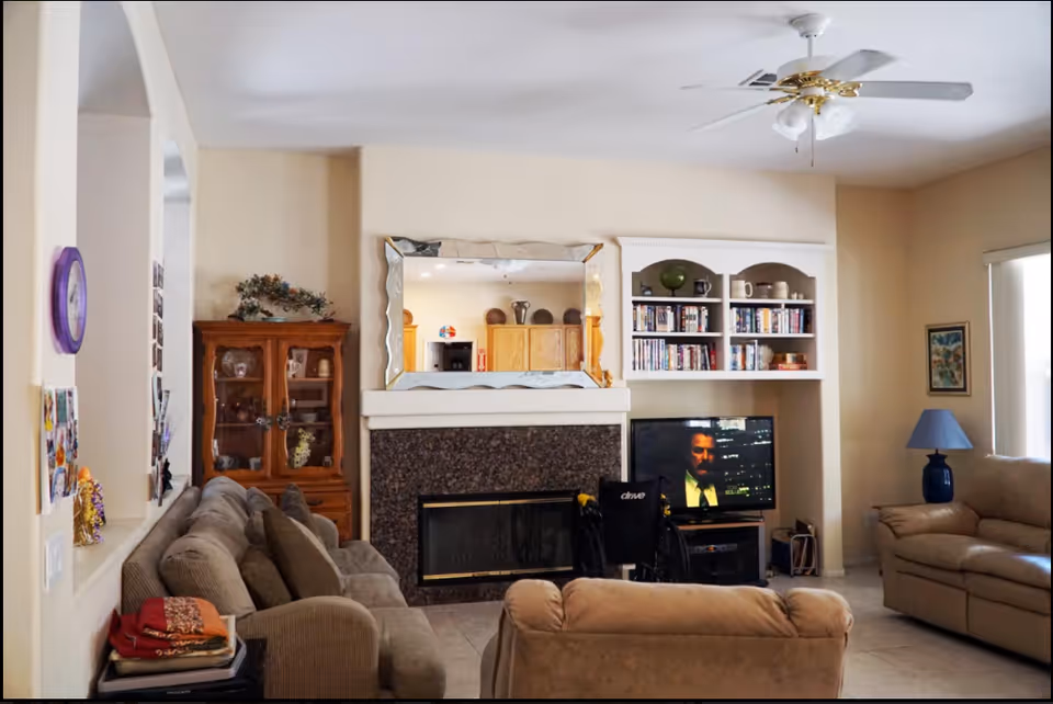 Living room of a senior care facility with sofas, a fireplace, shelving, and a wheelchair.