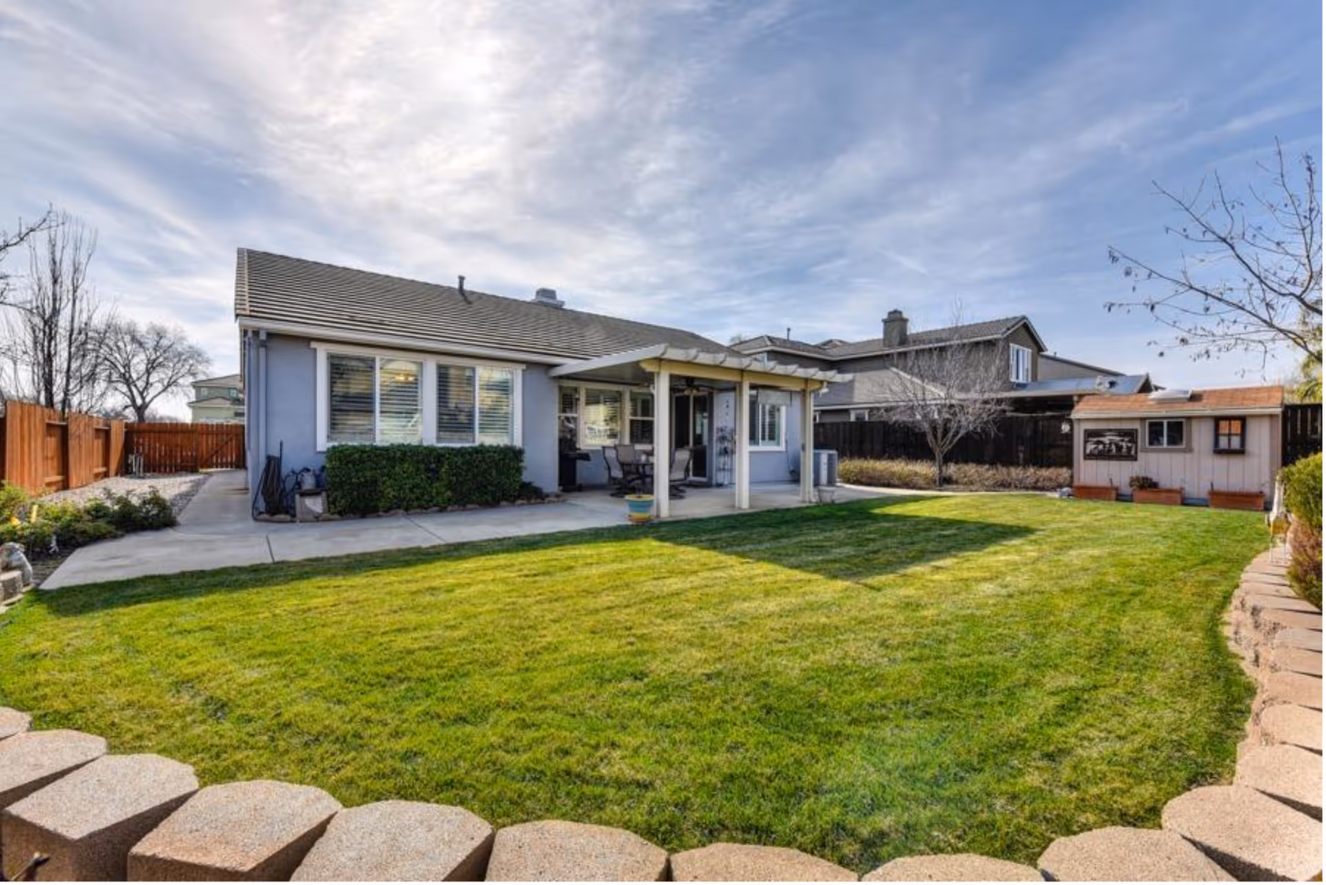 Backyard lawn with a patio, a single-story house, wooden fence, and shed.