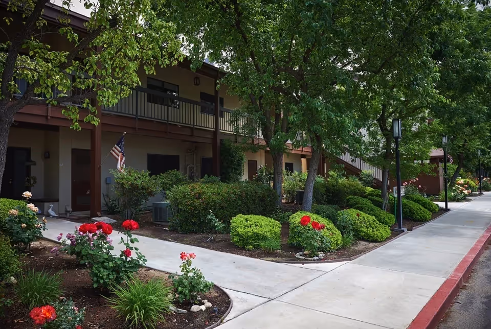 Landscaped sidewalk and flowerbeds leading past a two-story residential building with balconies and an American flag.