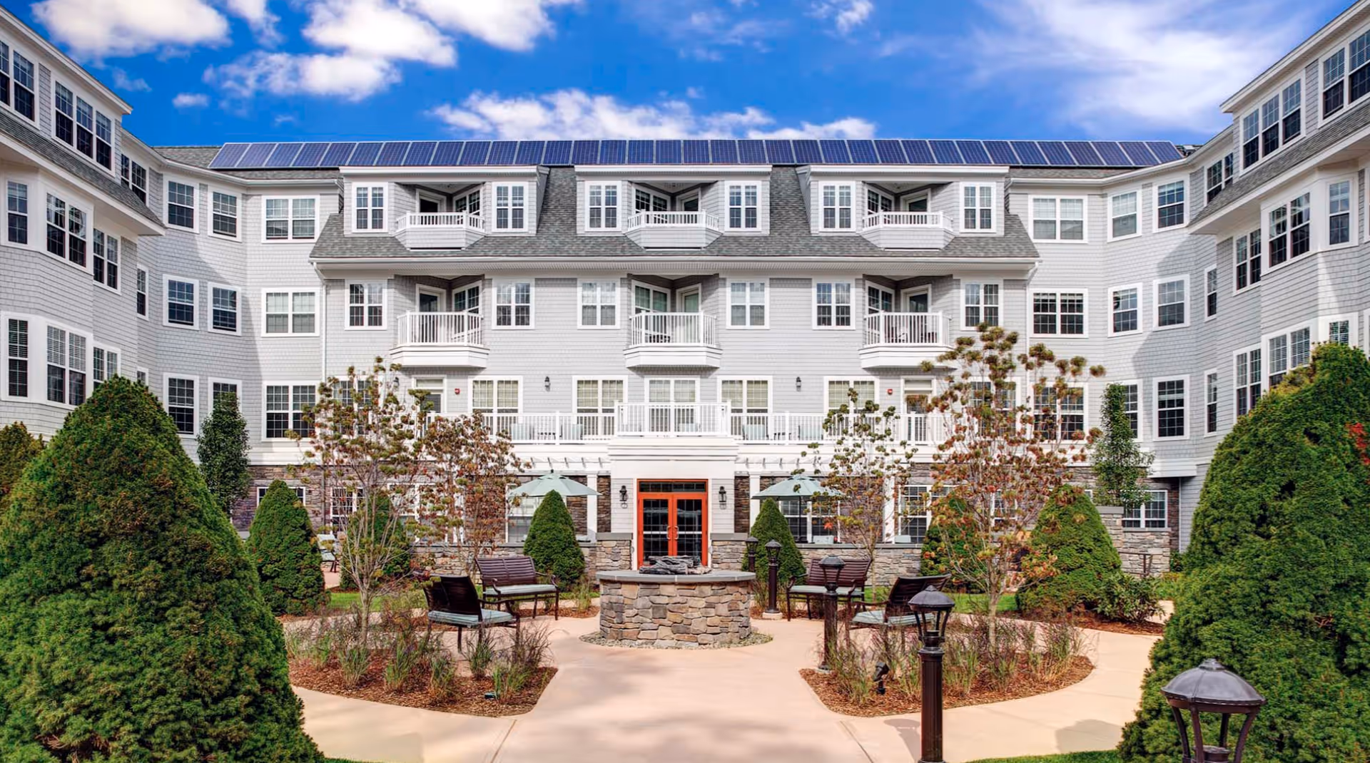 Exterior view of Atria Woodbriar Place, a multi-story senior living facility with white siding and numerous windows. The building features balconies and solar panels on the roof. In the foreground, there is a landscaped courtyard with trimmed bushes, trees, benches, umbrellas, and a stone fire pit under a blue sky with scattered clouds.
