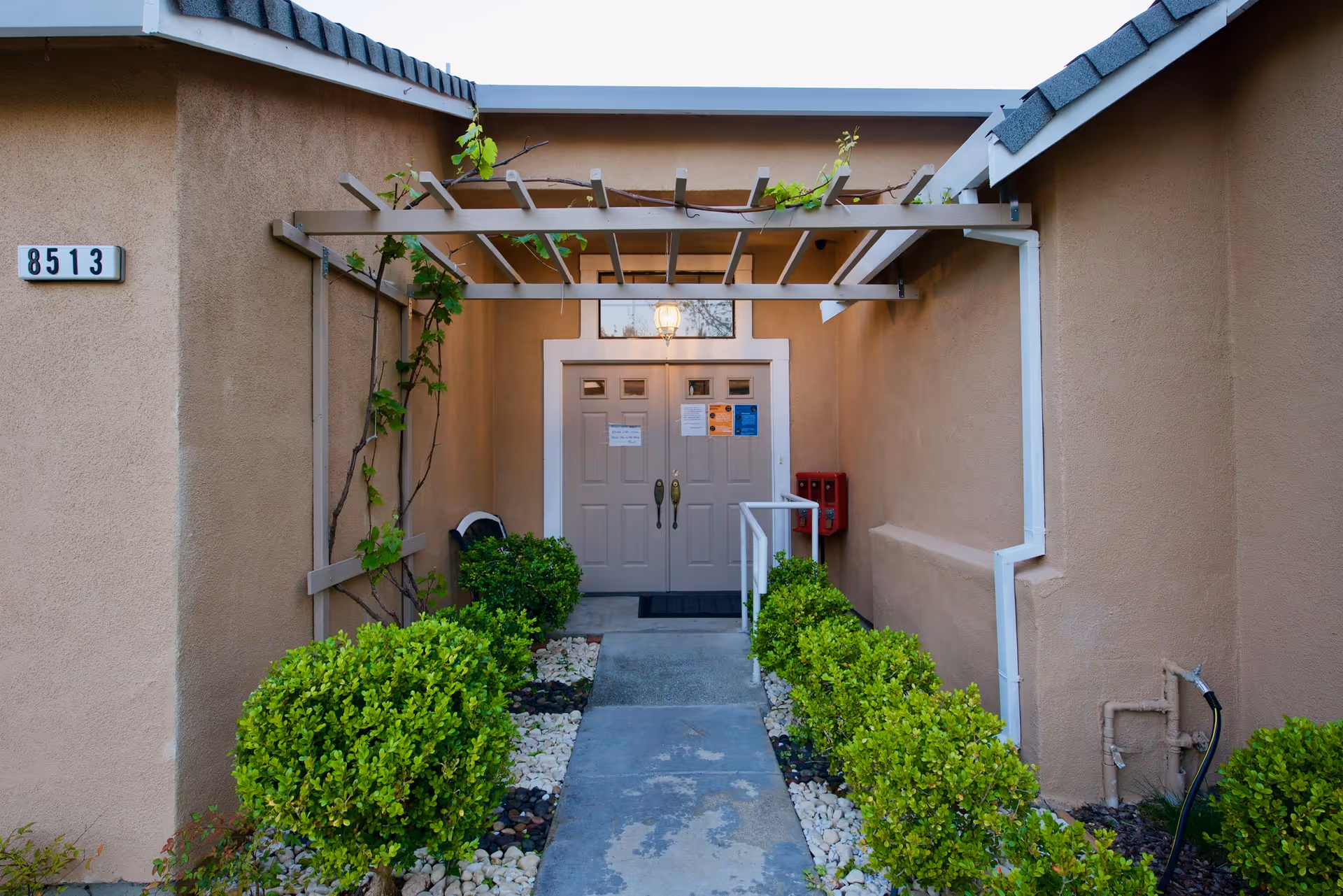 Beige exterior entrance with a walkway, climbing vines, and double doors.