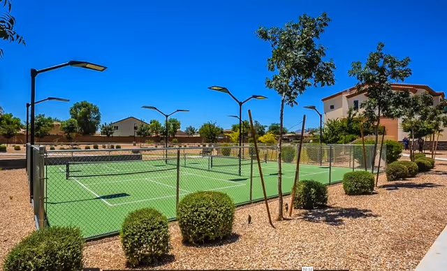 Outdoor tennis court surrounded by a chain-link fence with several small trees and bushes planted around it. There are tall street lamps around the court, and residential buildings are visible in the background under a clear blue sky.