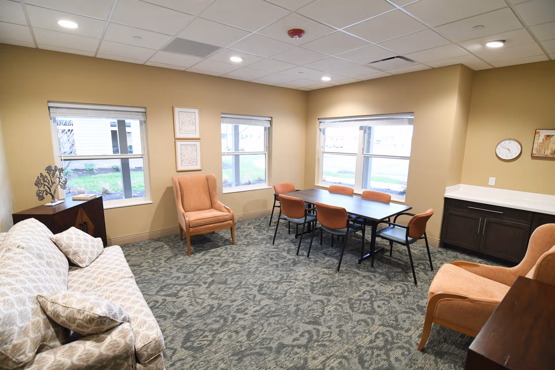 Common living room in a senior facility with sofa, chairs, and windows.
