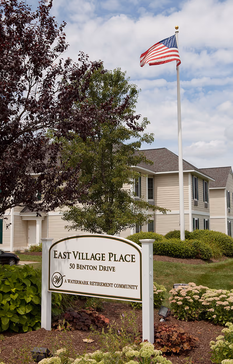 Exterior view of East Village Place with a front sign, trees, and American flag.